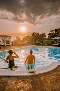 Rear view of couple in swimming pool against sky during sunset