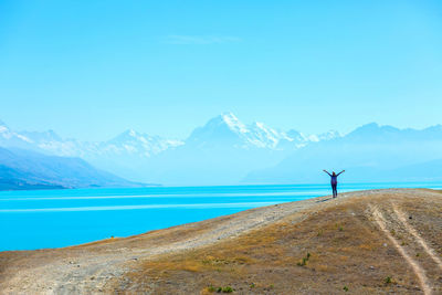 Woman with arms outstretched standing at lakeshore against mountains