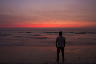 Rear view of silhouette man standing at beach against cloudy sky during sunset