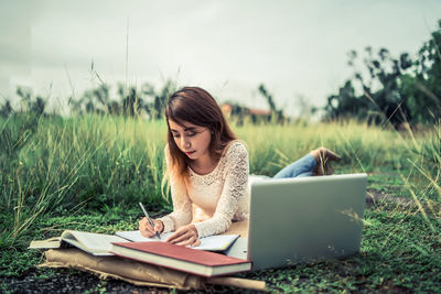 Young woman writing in book by laptop on field