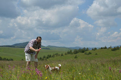 Man is playing with his dog on a meadow, surrounded with pink flowers and with hills in background