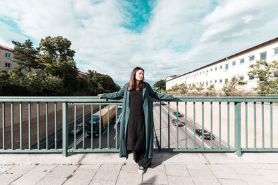 Mid adult woman standing by railing on footbridge against cloudy sky