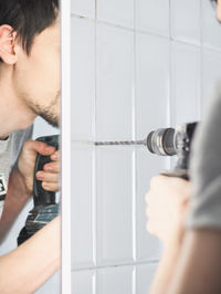 A young man drills between the seams on a tiled wall.