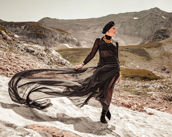 A stylish strong woman in a black dress fluttering in the wind walks on the snowy mountains