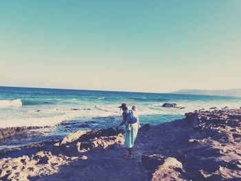 Mother piggybacking child on rock at beach against clear sky