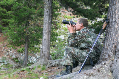 Man holding binoculars while sitting against tree trunk in forest