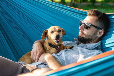 Handsome european man in sunglasses is resting in hammock with his cute little dog.