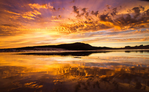 Scenic view of seascape by mountain against cloudy sky during sunset