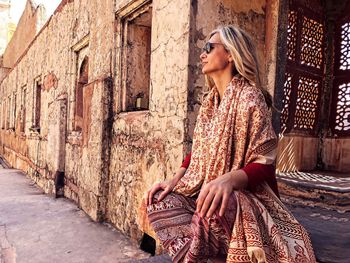 Mature woman wearing sunglasses and traditional clothing sitting against old building