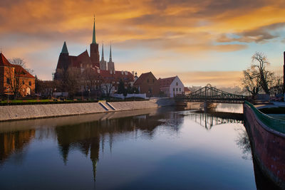 Bridge over river by buildings against sky during sunset