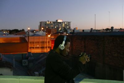 Side view of man wearing headphones while photographing through digital camera in city