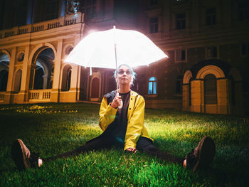 Young woman with illuminated umbrella sitting on grassy field at night