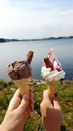 Cropped hands of female friends having ice cream cones against lake