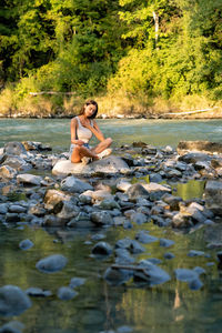 Beautiful young woman sitting on stone in the mountain river with closed eyes breathing 