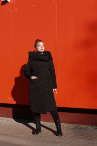 Fashionable teenage girl wearing black warm clothing standing against red wall during sunny day
