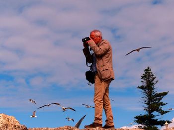 Side view of man photographing with camera while standing on field against cloudy sky