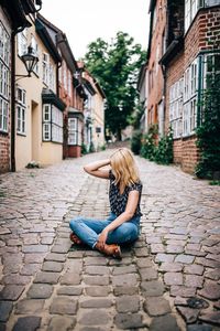 Full length of blond woman sitting on cobbled footpath amidst houses