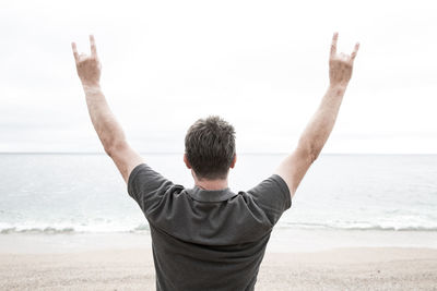 Rear view of man showing rock sign while standing at beach