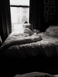 Rear view of shirtless man sitting on bed against window at home