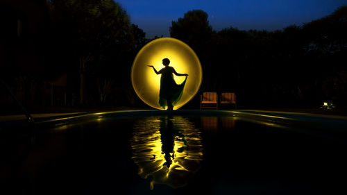 Silhouette woman standing by illuminated lighting equipment reflecting on swimming pool at night