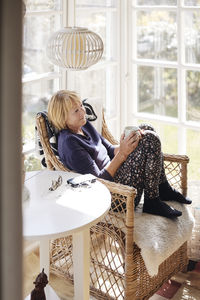 Senior woman with coffee cup looking away while relaxing on chair at home