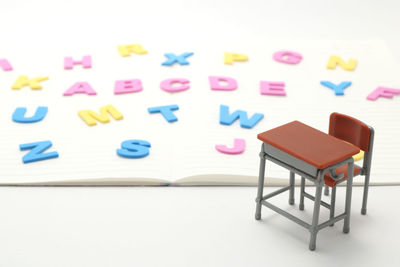 Close-up of miniature desk and chair with multi colored alphabets on book over white background