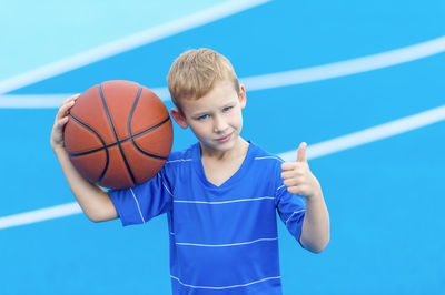 Portrait of boy gesturing and holding basketball while standing on sports court