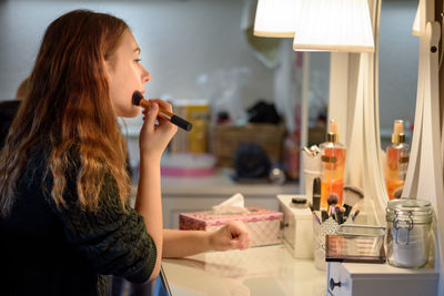 Side view of woman applying make-up in front of mirror at home