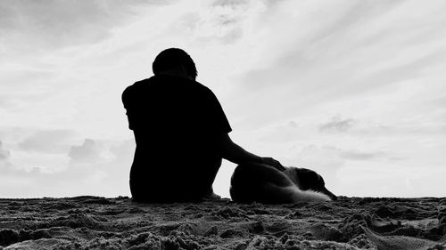 Rear view of man with dog sitting at beach against cloudy sky