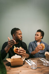 Male friends talking to each other while eating burgers sitting at restaurant