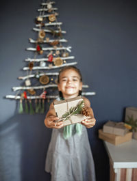 Portrait of smiling girl holding gift against christmas tree hanging on wall