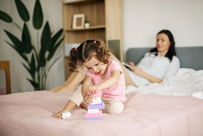 A cute girl is playing with a constructor on the bed while her mom is working on a laptop