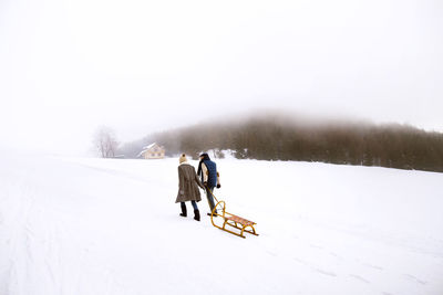 Back view of senior couple with sledge walking side by side in snow-covered landscape