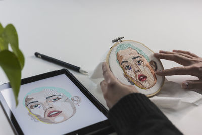 Young female freelancer holding embroidery frame by digital tablet at studio