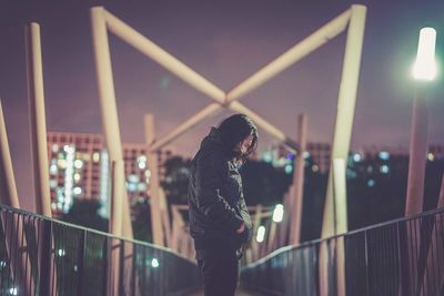 Side view of man standing on footbridge against illuminated city at night