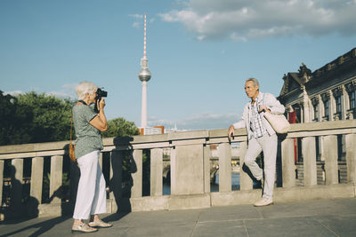 Full length of woman taking photograph of senior man while standing on bridge against tower in city