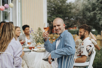 Side view portrait of smiling young man holding wineglass while sitting on chair with friends during dinner party at caf