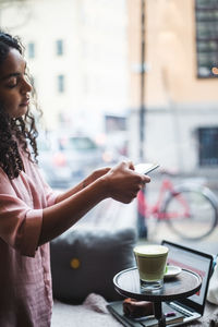 Young female blogger photographing tea and snack on table through smart phone at creative office