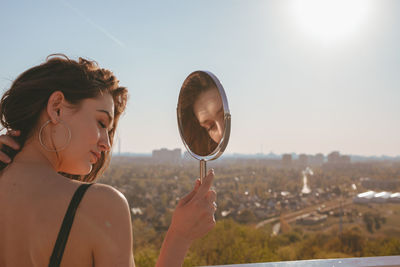 Rear view of young woman with mirror standing against clear sky