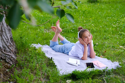 Teen girl lies listens to music in headphones on the grass in the park with books and notebooks