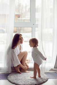 Mother in a white robe sits with a child a blonde daughter at a large window of the house person