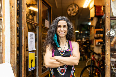 Positive adult ethnic female mechanic in colorful apron with protective mask smiling and looking at camera with arms crossed standing on entrance of bike service workshop