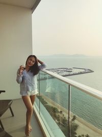 Portrait of smiling woman with wineglass standing at balcony by sea