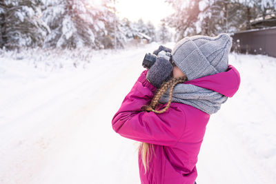 Girl in winter clothes is taking photo, standing sideways on road against snowy pine trees. 