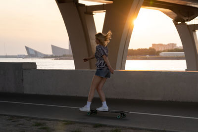 Active woman enjoying summer evening skating on longboard along river with city and sunset view