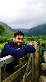 Portrait of smiling man leaning over fence on mountains against cloudy sky