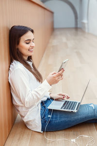 Smiling young woman using phone while sitting on floor