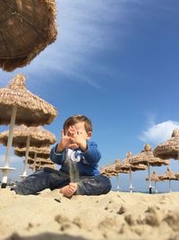 Surface level of boy playing with sand by thatched roof parasols against sky