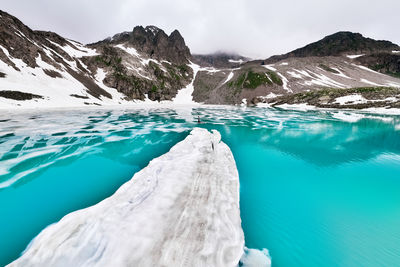 A man swims on a paddleboard in a mountain turquoise lake partially covered with snow, surrounded by