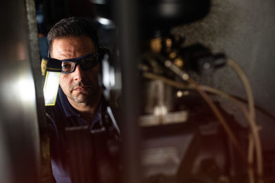 Professional male mechanic in protective glasses and headphones using flashlight while checking details of machine during repair works in workshop